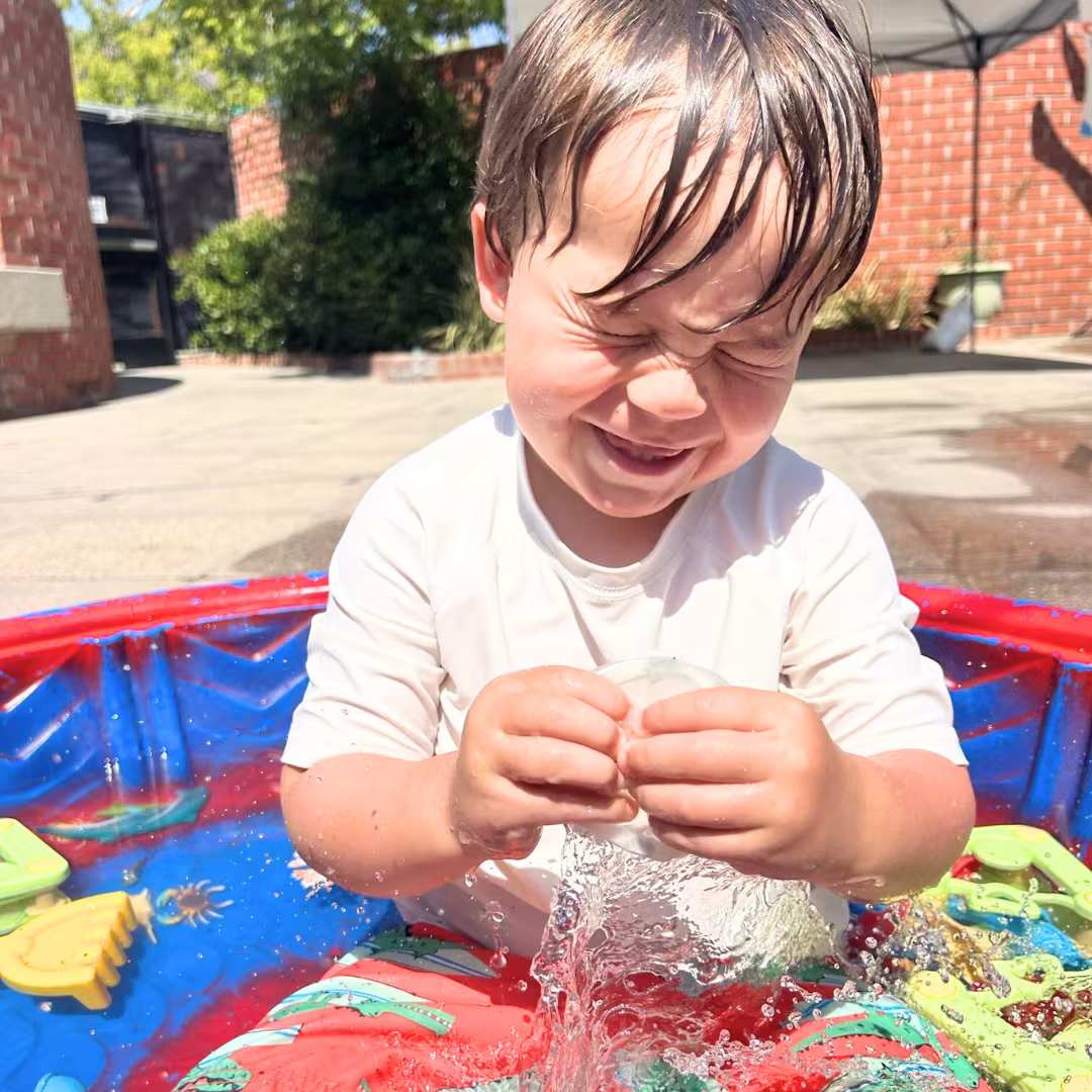 Young child with wet hair playing and splashing water in a small blue and red plastic pool outdoors on a sunny day.