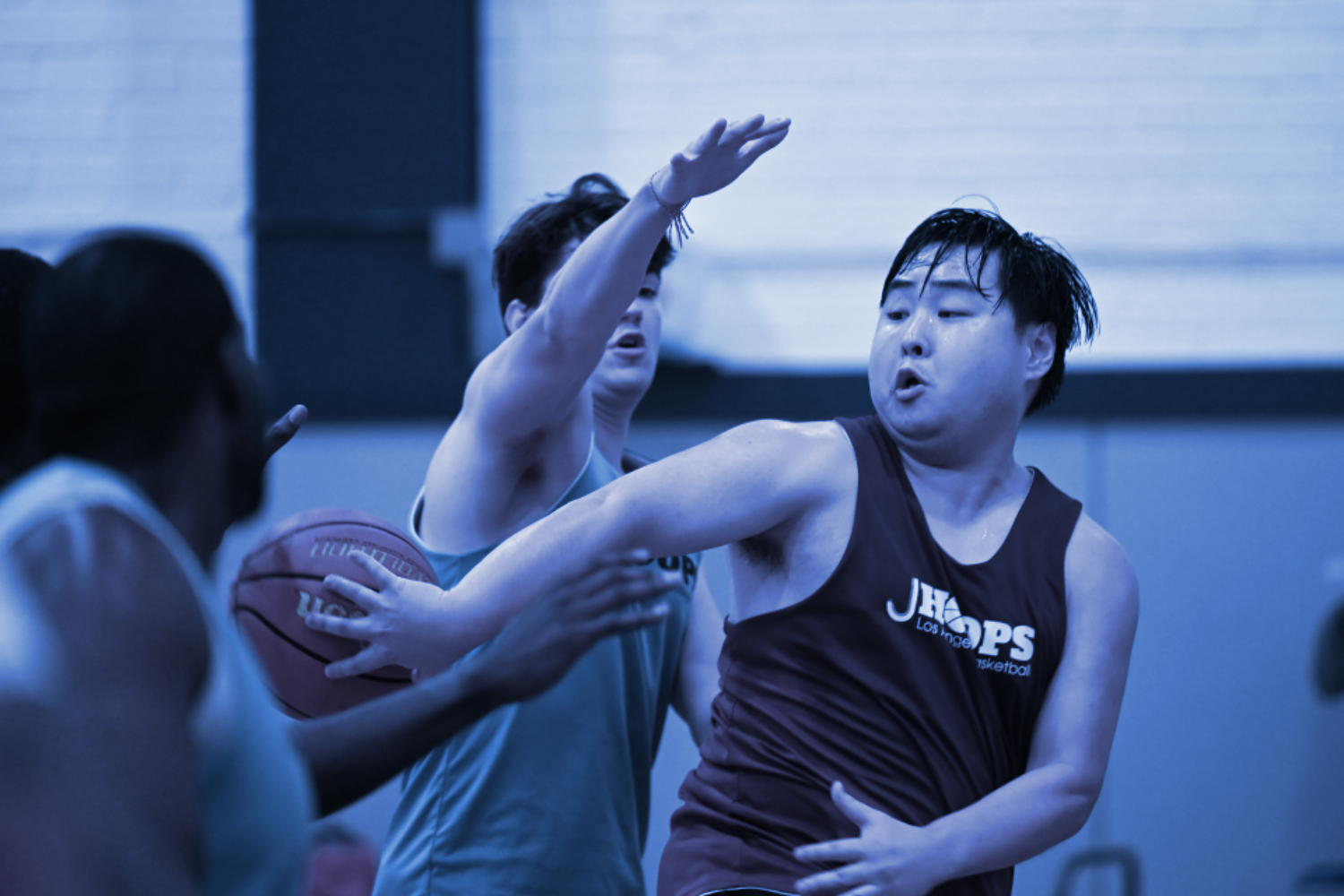 A basketball player in a black jersey leaps to shoot a basketball while another player in a gray shirt attempts to block the shot. They are on a court inside the Jewish Community Center in Mid City LA, part of the Adult Basketball League.