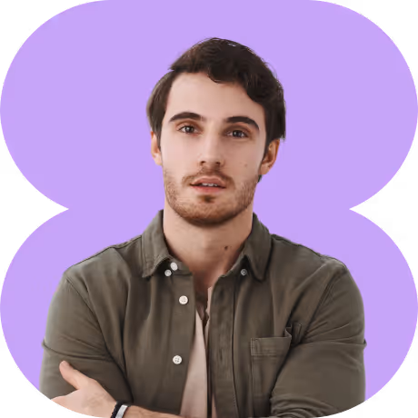 Young man with dark hair, light facial hair, wearing an olive green button-up shirt over a beige t-shirt, standing with arms crossed against a purple background.