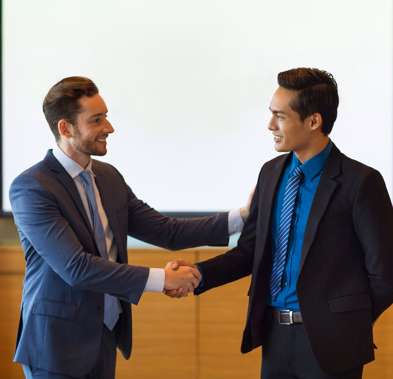 Two businessmen dressed in suits smiling and shaking hands in an office setting.