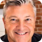 Smiling middle-aged man with gray hair and blue eyes in front of a brick wall.