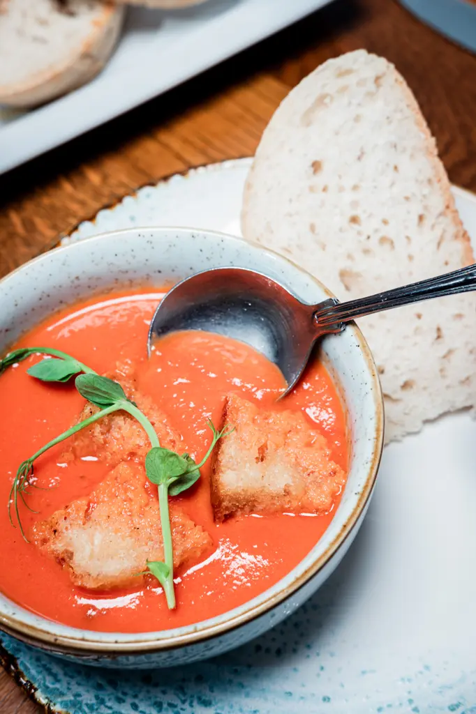 A closeup image of tomato soup with some white bread both off to the side of the bowl and submerged in the soup.