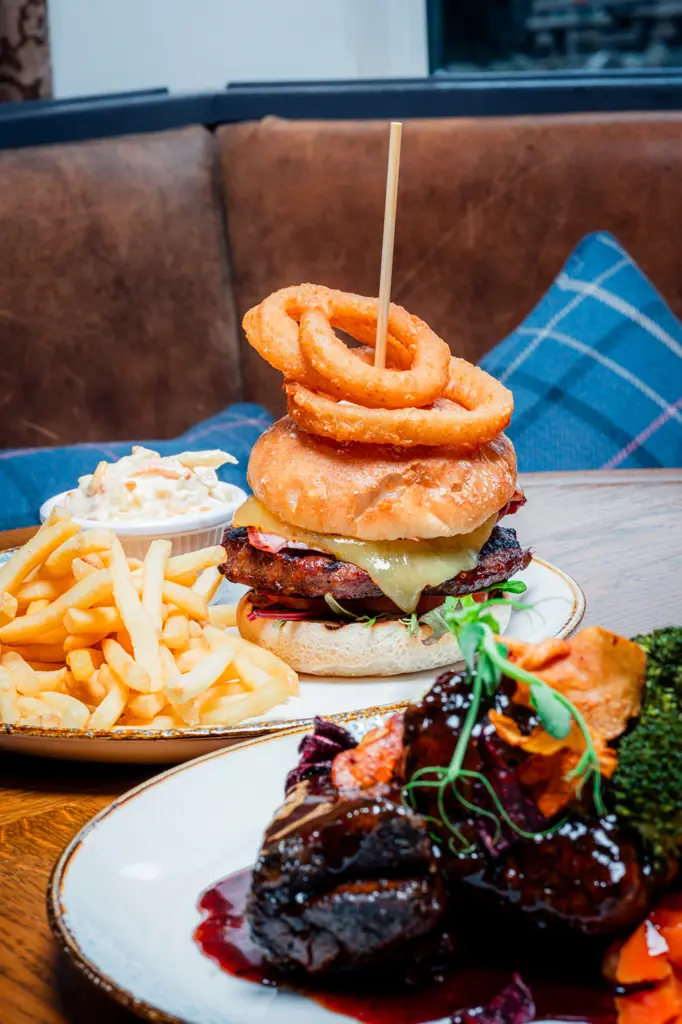 A classic cheeseburger with onion rings on top surrounded by coleslaw and a plate of chips on the side.