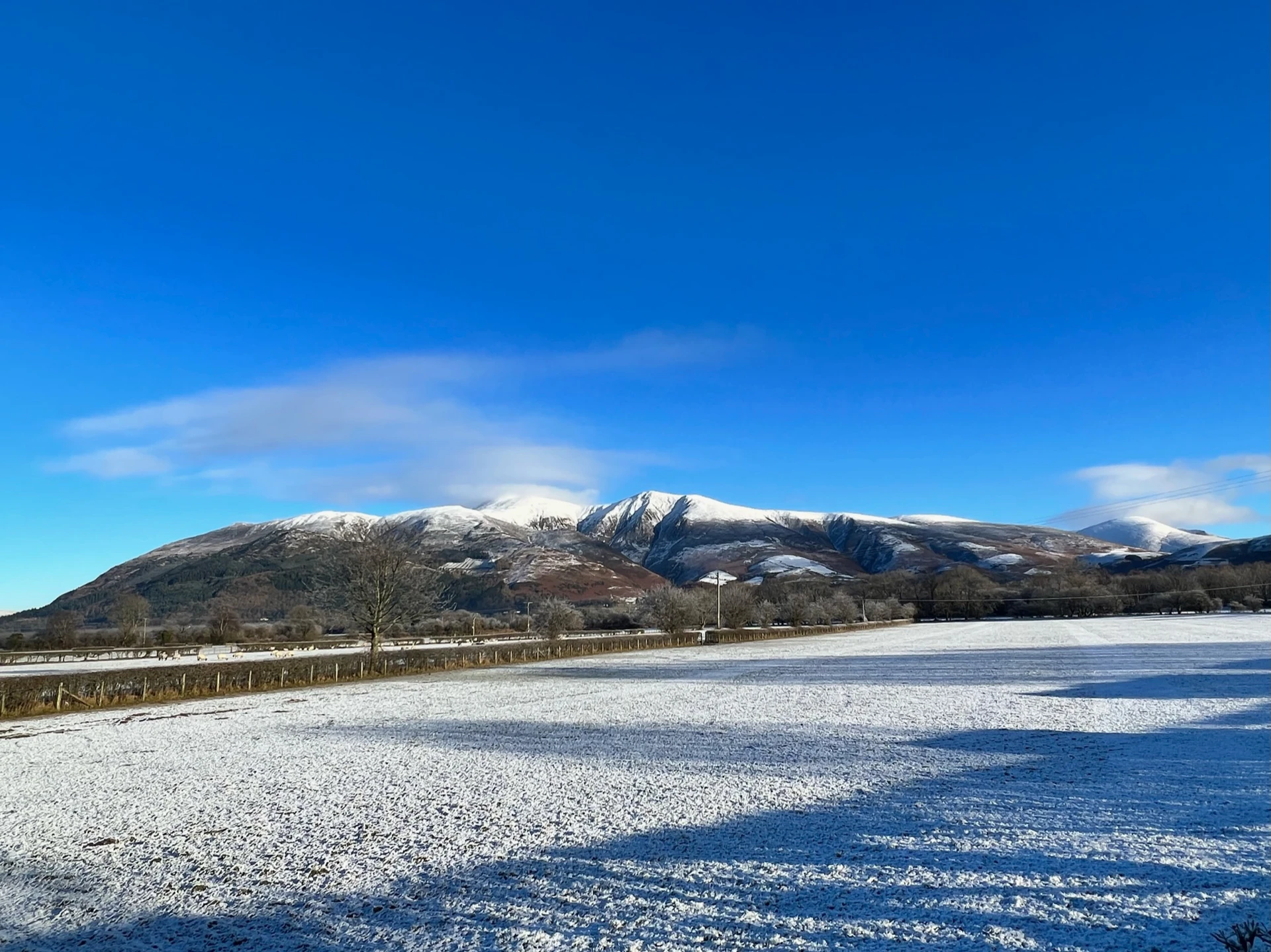 A snowy landscape in the Lake District with a bright sunny sky and rolling snowy mountains in the distance.