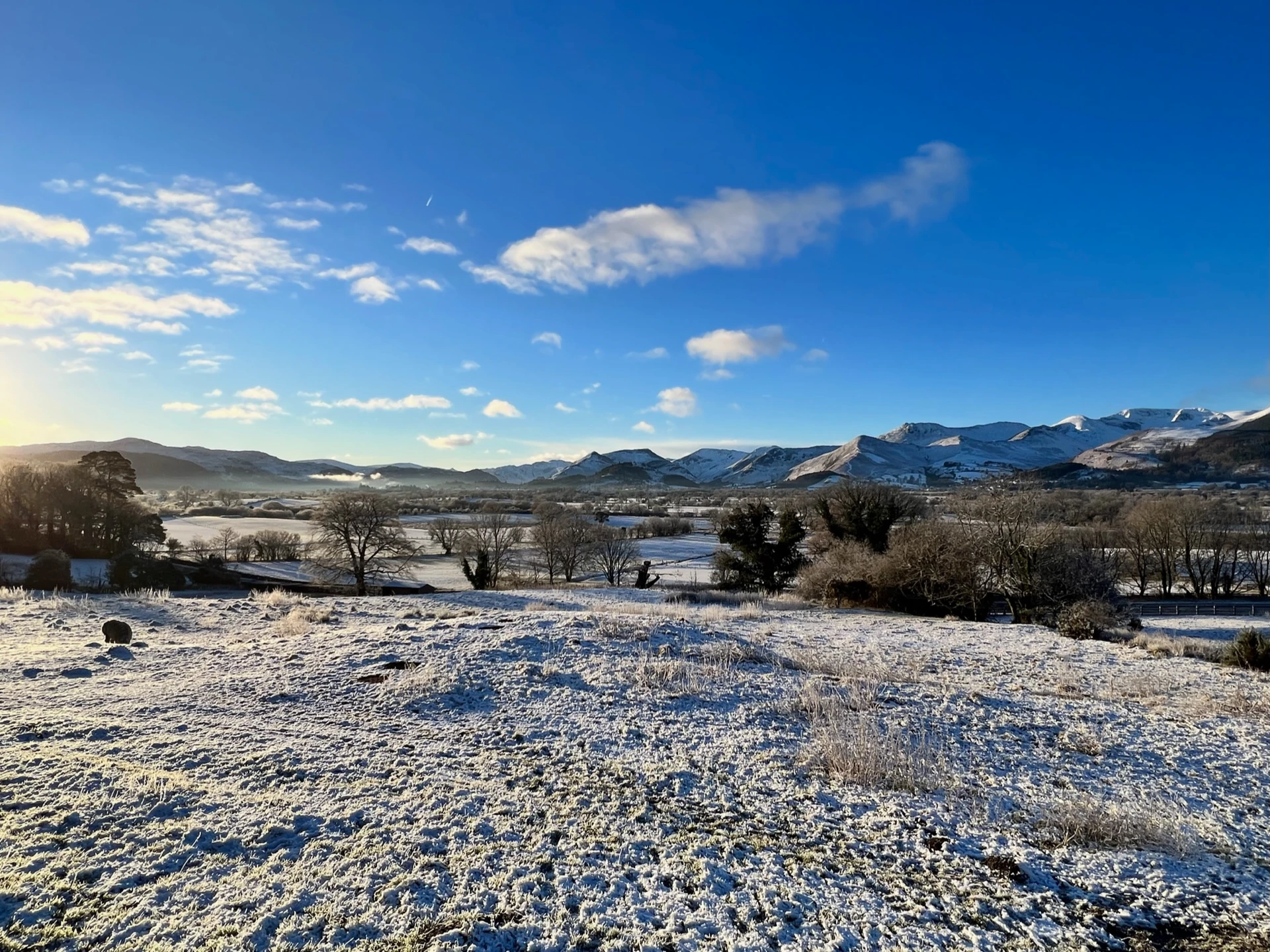 A snowy landscape in the Lake District with a bright sunny sky and rolling snowy mountains in the distance.