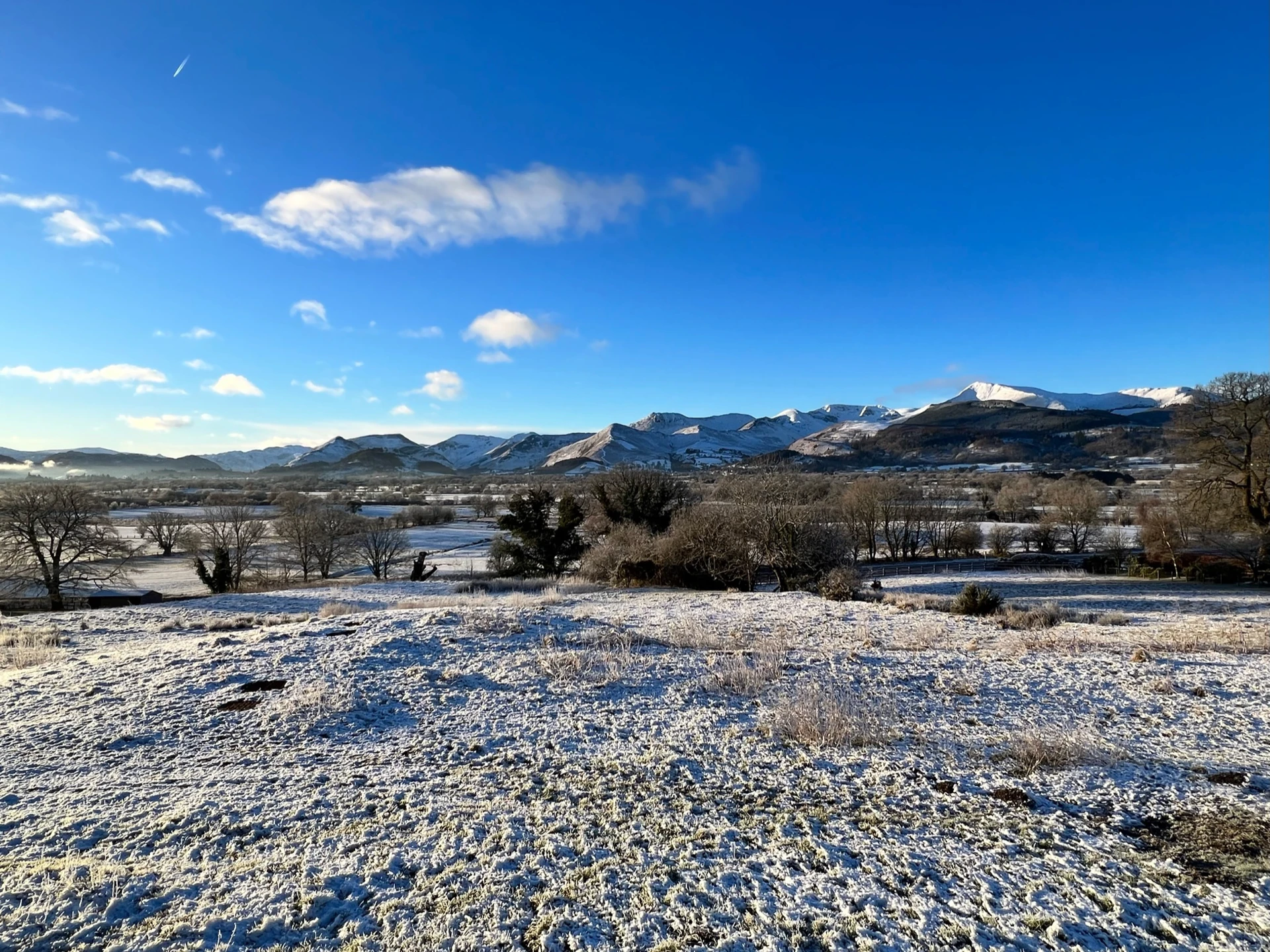 A snowy landscape in the Lake District with a bright sunny sky and rolling snowy mountains in the distance.
