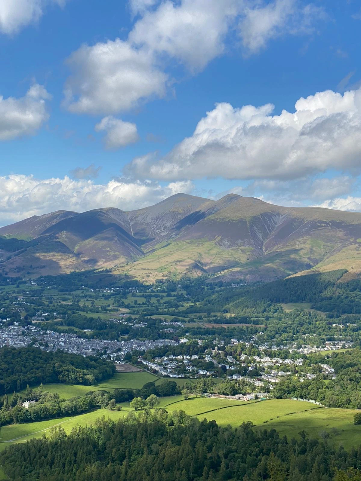 Rolling lush hills in the Lake District around Braithwaite Village under a sunny blue sky.