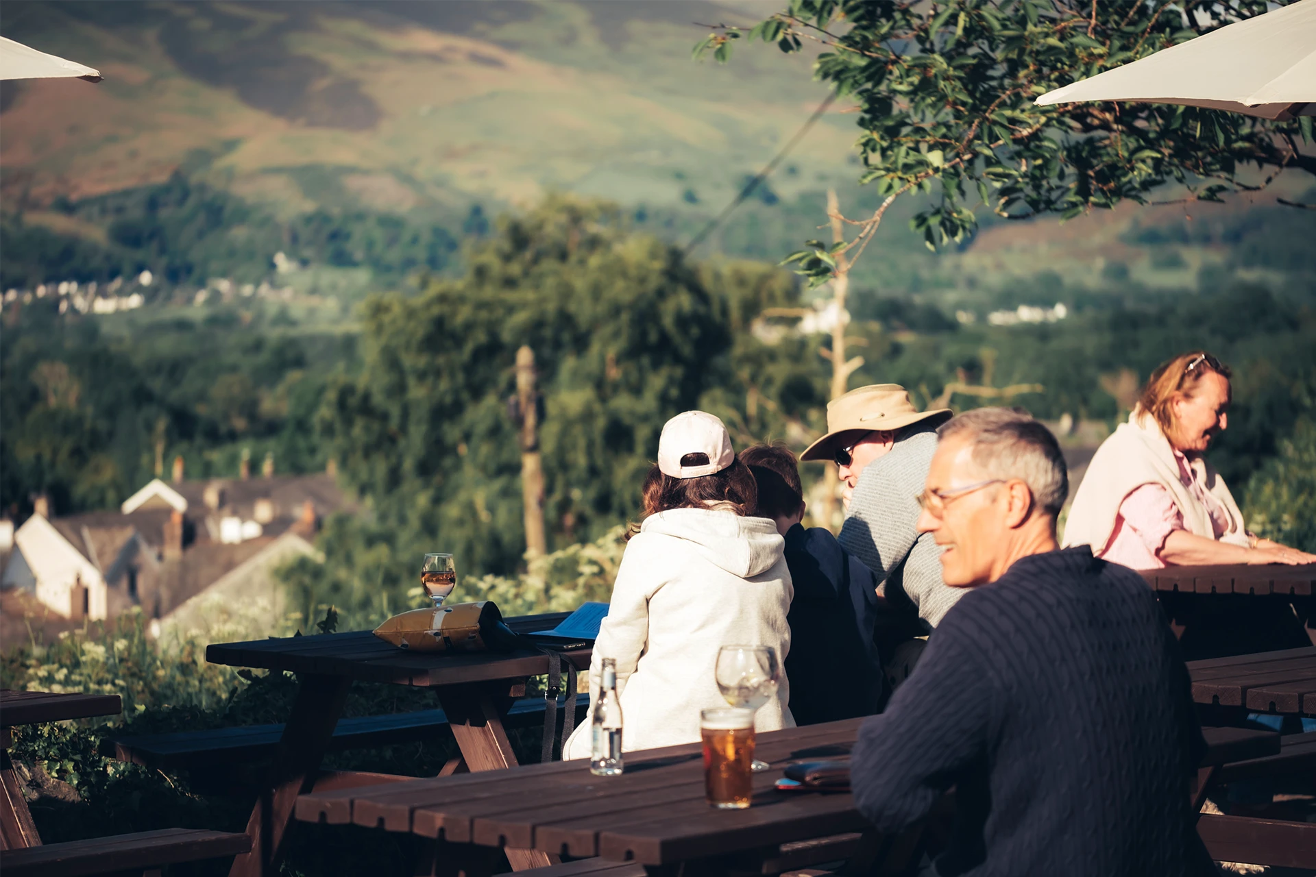 People sitting at outdoor wooden tables under umbrellas, enjoying a scenic view of rolling hills and greenery in the background. A few wear casual clothing and hats.
