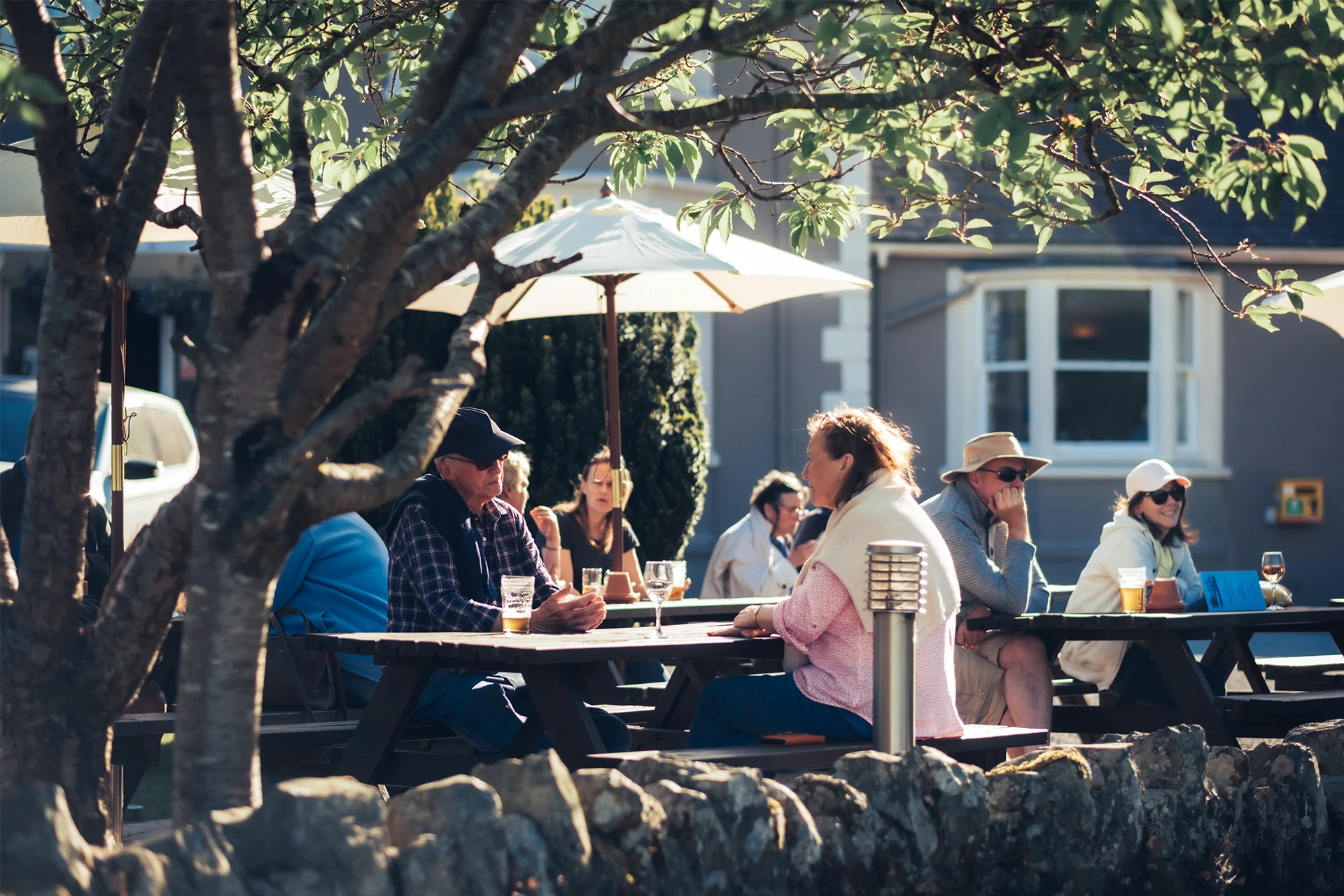 People sitting around picnic tables outdoors under trees, enjoying conversation and drinks. Some people are wearing hats and sunglasses. The setting is shaded by trees, with a building and an umbrella in the background.