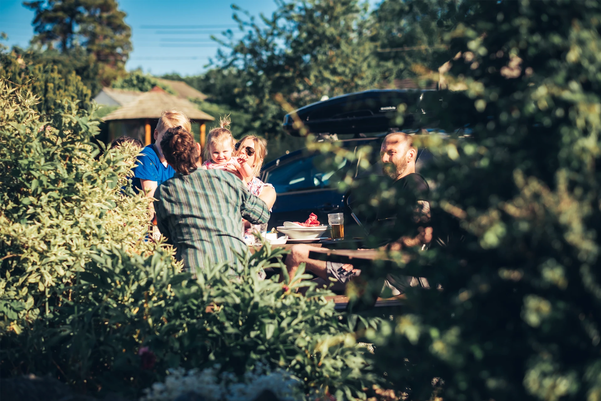 Four people sit together at a table outdoors, surrounded by lush greenery and plants. They appear to be engaged in conversation under the shade of an umbrella on a sunny day.