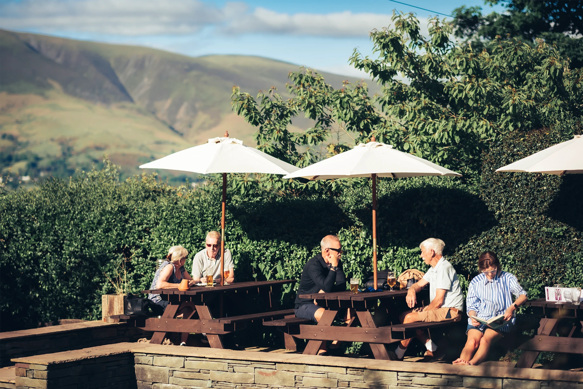 A group of people sitting at wooden picnic tables under white umbrellas, surrounded by lush greenery and hills in the background. The setting appears to be a scenic outdoor area with a relaxed, social atmosphere.