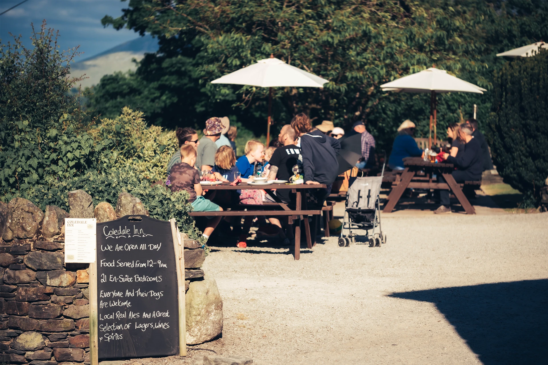 Groups of people sitting at picnic tables with umbrellas on a patio surrounded by greenery. A chalkboard menu is visible in the foreground. It appears to be a sunny day.