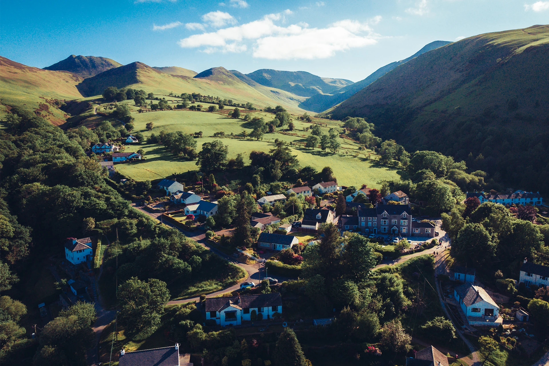 Aerial view of a small village nestled in a lush, green valley surrounded by hills. The landscape features scattered houses, winding roads, and patches of trees under a bright blue sky with scattered clouds.