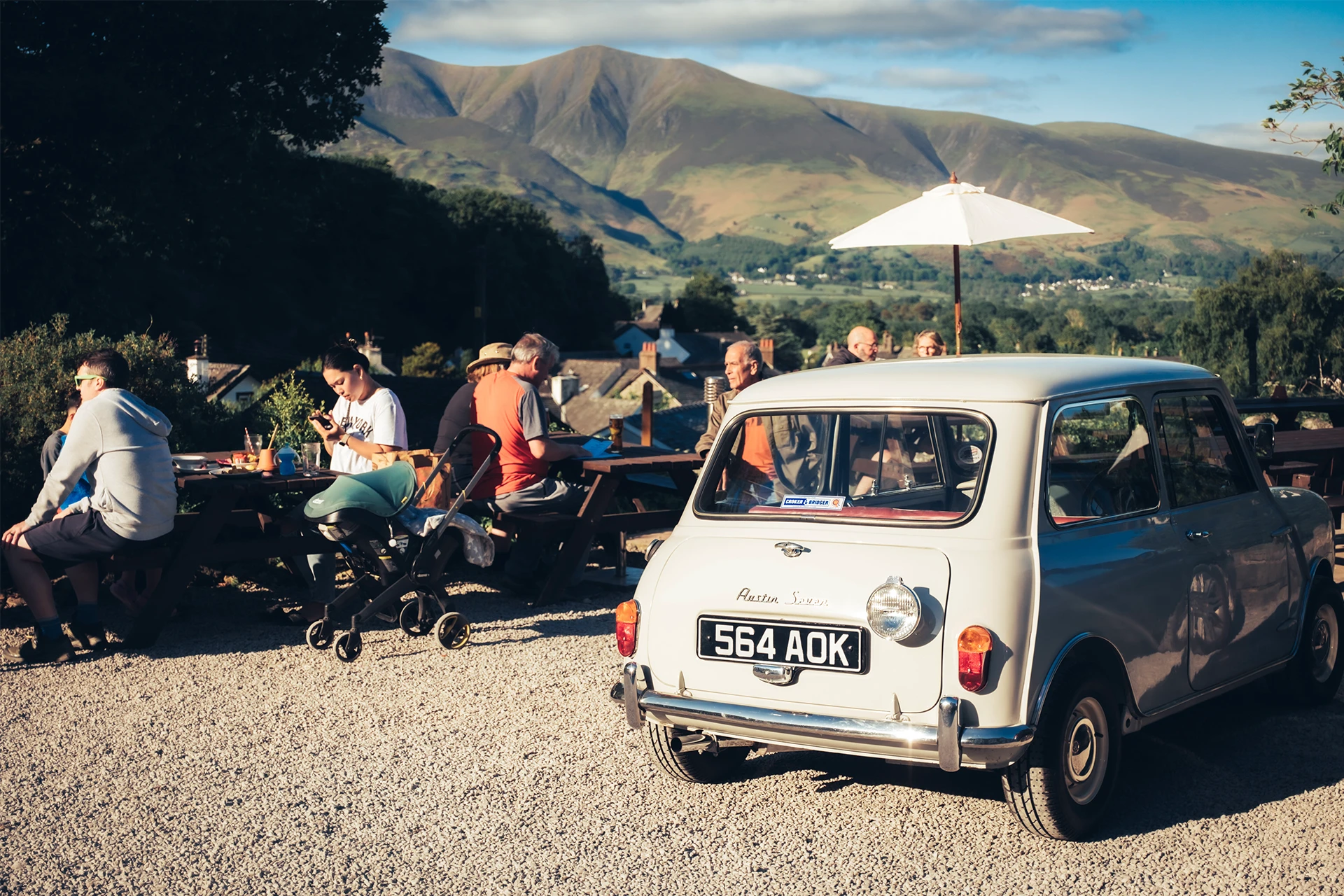 A vintage white car is parked on a gravel surface in the foreground. People are seated at outdoor picnic tables under a large white umbrella. Green hills and a mountain range are visible in the background under a blue sky.