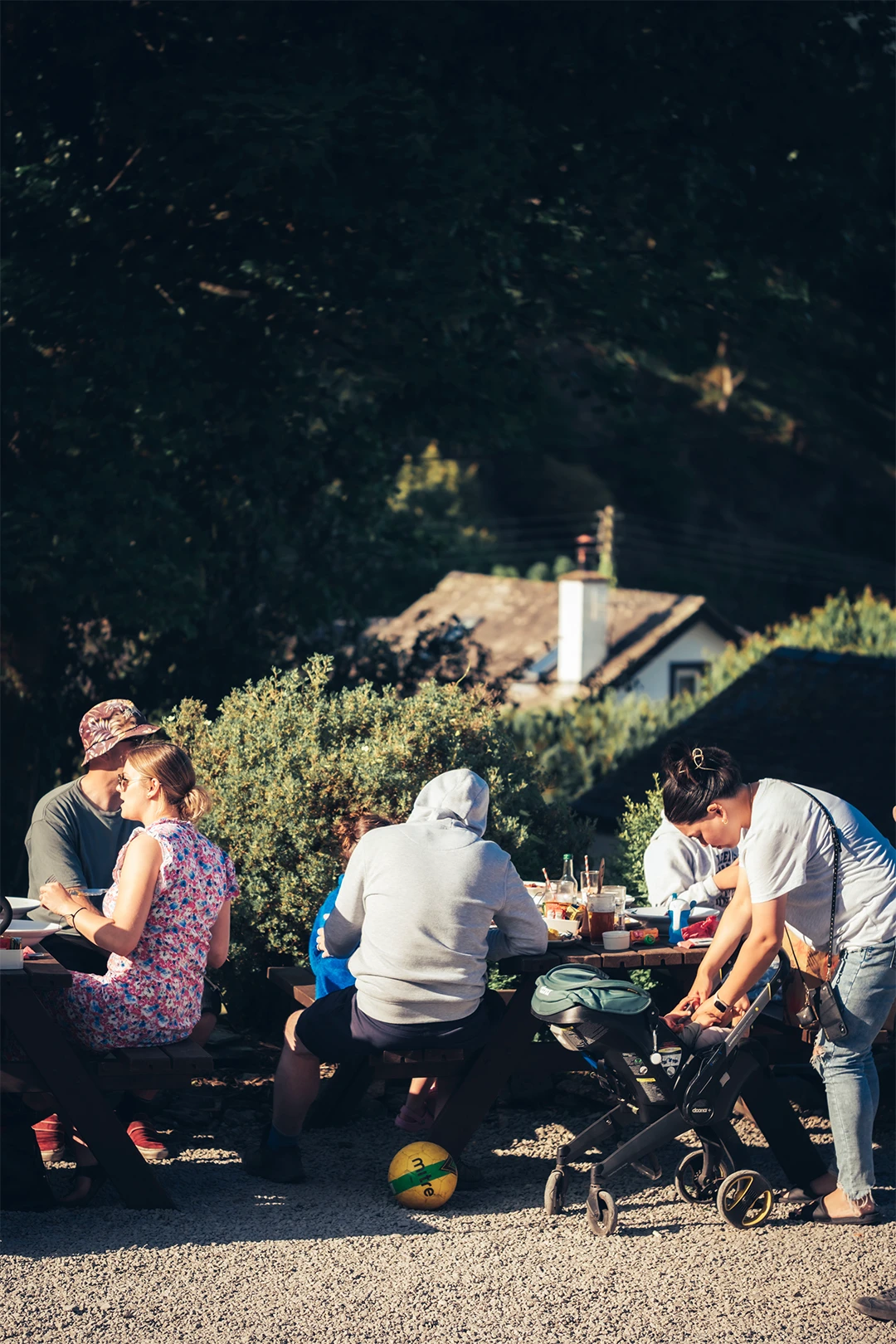 A group of people sitting around a picnic table outdoors, surrounded by greenery and dappled sunlight. Some are eating, and others are engaged in conversation. A stroller is visible beside the table.