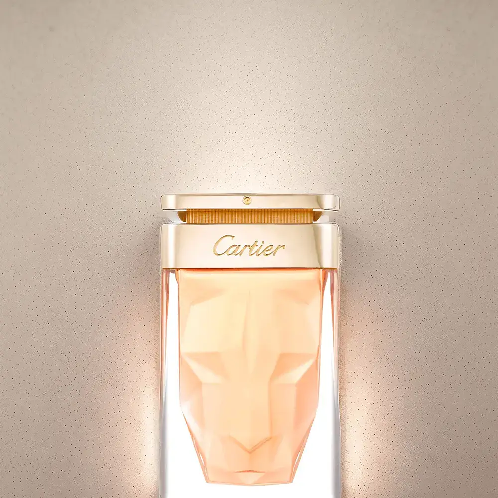 Cartier perfume in a geometric faceted bottle with a gold cap against a neutral beige background. The amber/peach-colored fragrance has a sculptural quality with dramatic lighting that enhances its luxurious appearance and faceted glass design.