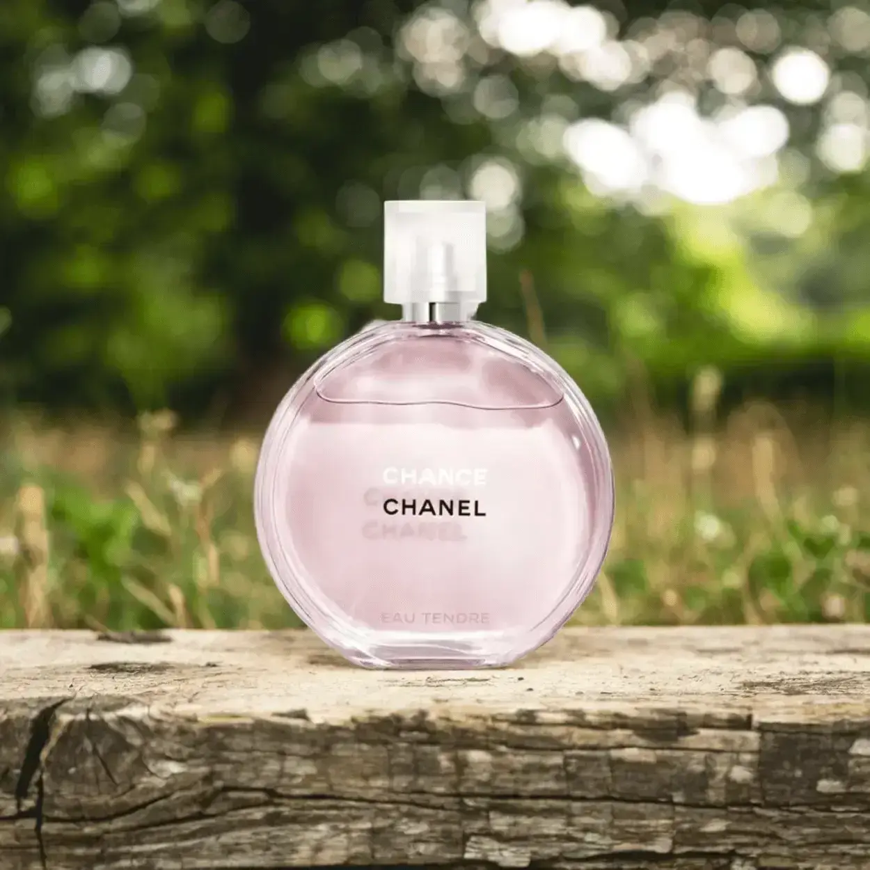 Chanel Chance Eau Tendre perfume bottle set in a natural outdoor environment. The round, light pink fragrance bottle is positioned on a rustic wooden surface against a blurred green background of grass or plants, creating an elegant yet natural aesthetic.