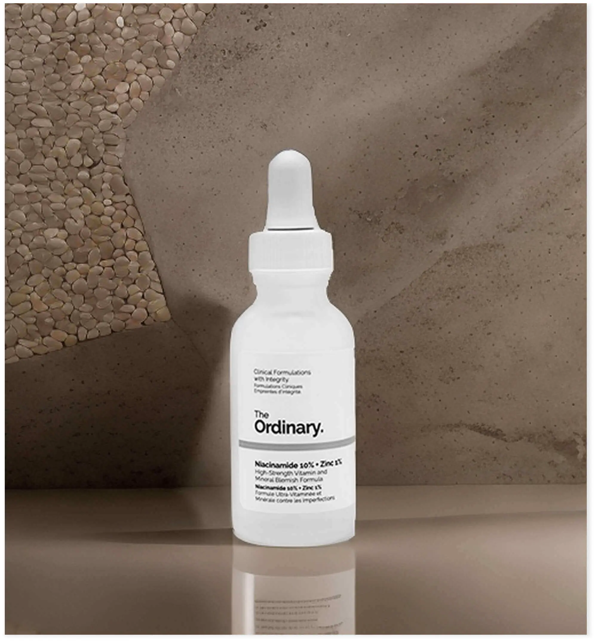 The Ordinary skincare serum on a textured concrete surface with pebble accents. The minimalist white dropper bottle with black text showcases the brand's clinical, science-focused aesthetic with clean lines and pharmaceutical-style packaging.