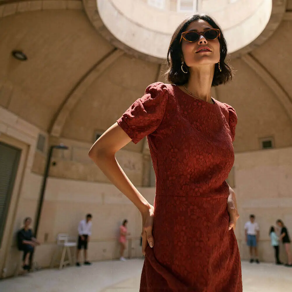 Fashion photography featuring a woman in a textured rust-red dress wearing cat-eye sunglasses in an architectural setting with a domed ceiling. The upward angle creates a dramatic composition highlighting both the garment and the model's accessories.