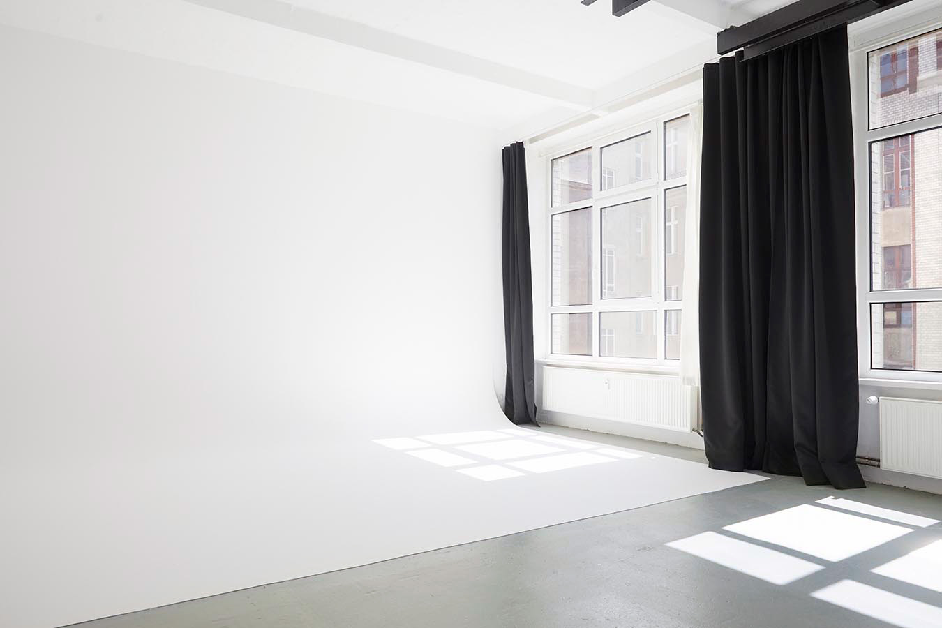 Daylight photo studio for rent in Berlin Neukoelln with a white infinity curve wall and large south-facing windows with black blackout curtains.