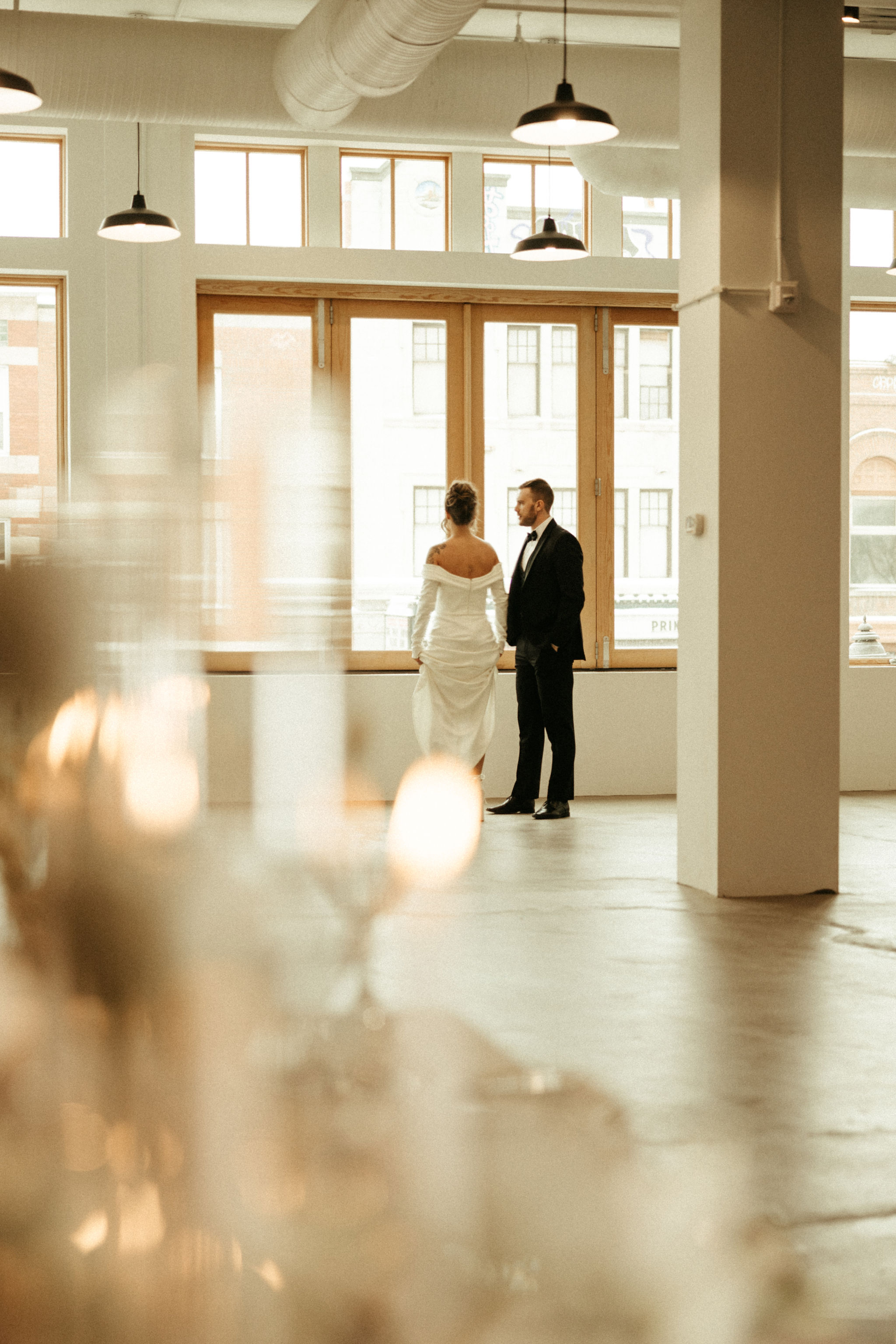 Bride in white gown and groom in black tuxedo standing side by side facing large Whyte Avenue windows in a bright, modern room.