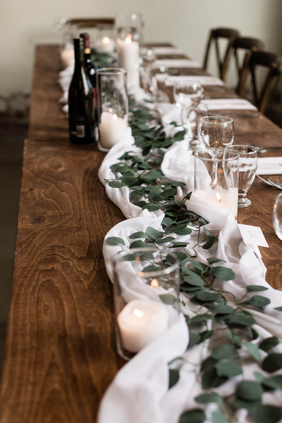 Long wooden farmhouse table with white fabric and greenery centerpiece, glass candle holders with lit candles, wine bottles, and neatly arranged glassware and place cards.