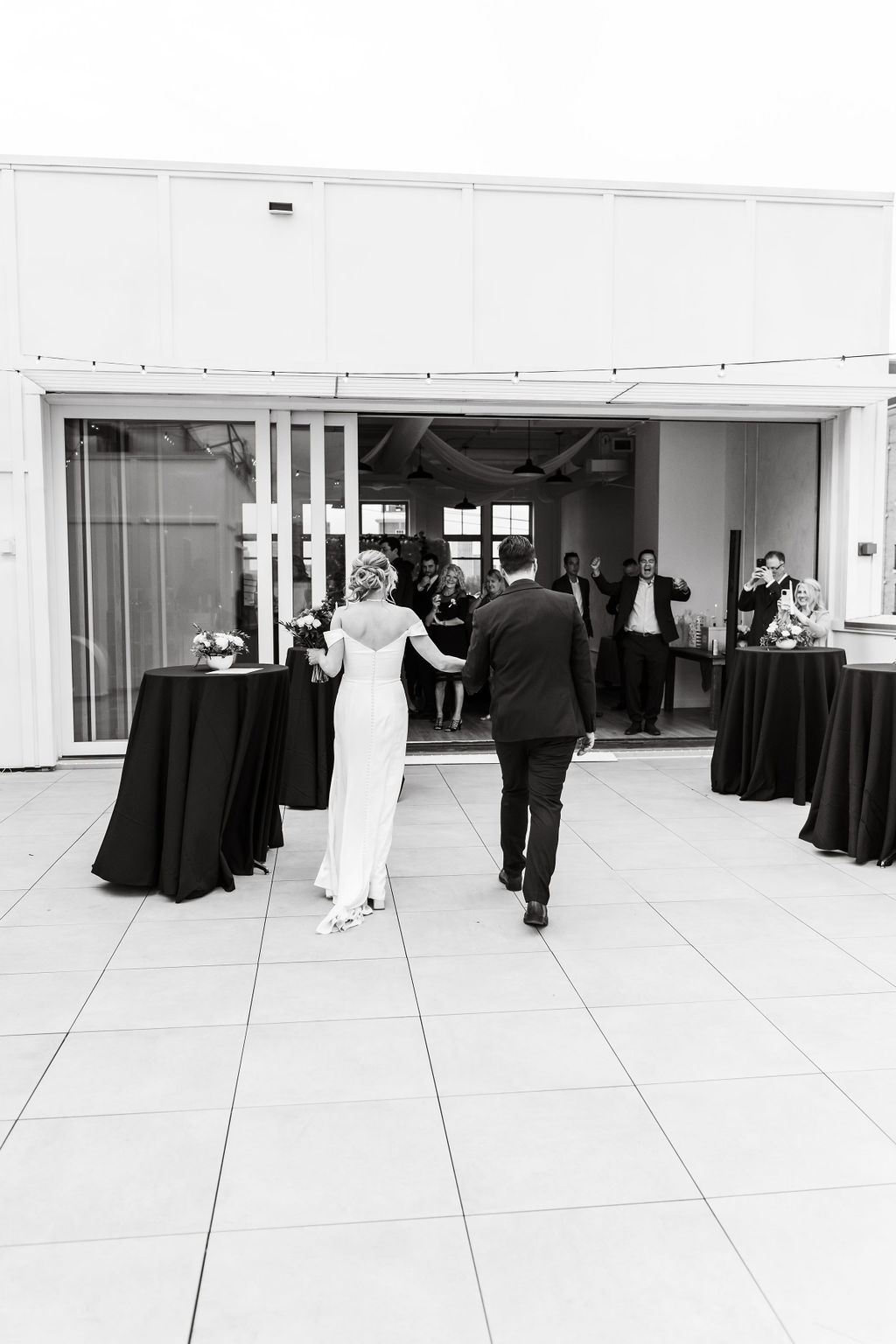 Bride and groom holding hands walking into a reception area with guests cheering and taking photos.