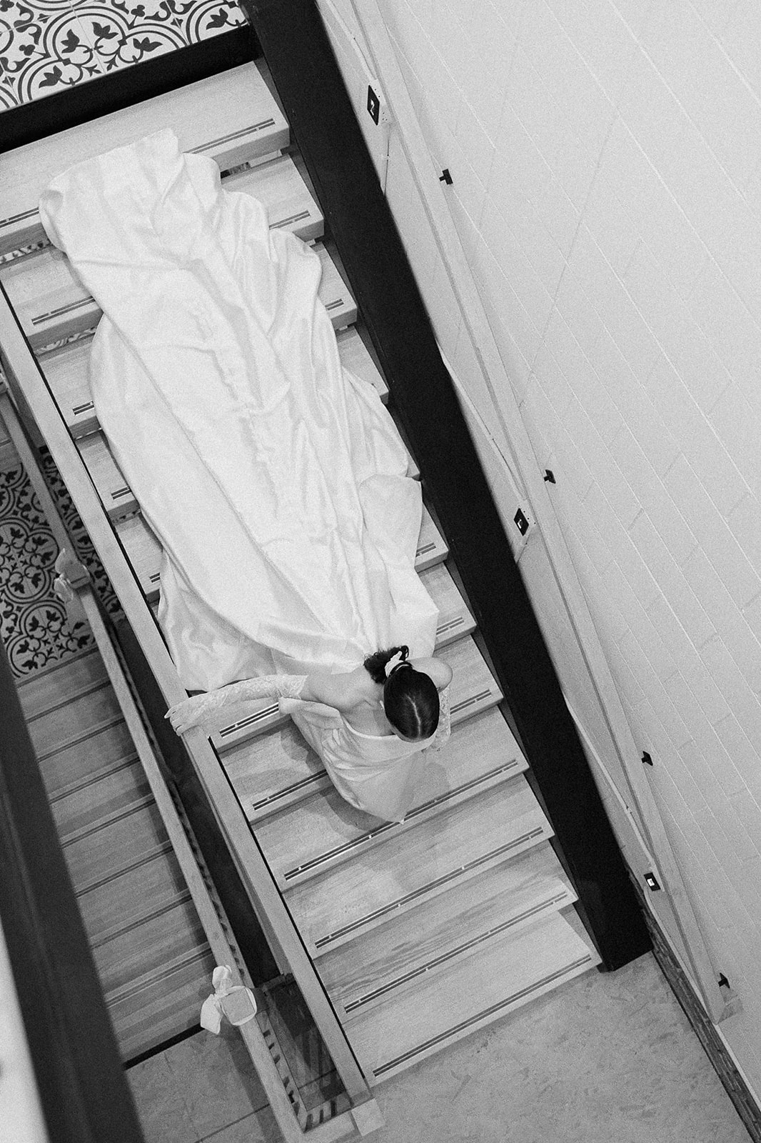 Overhead black and white photo of a bride descending a staircase in a flowing wedding gown and long lace gloves.