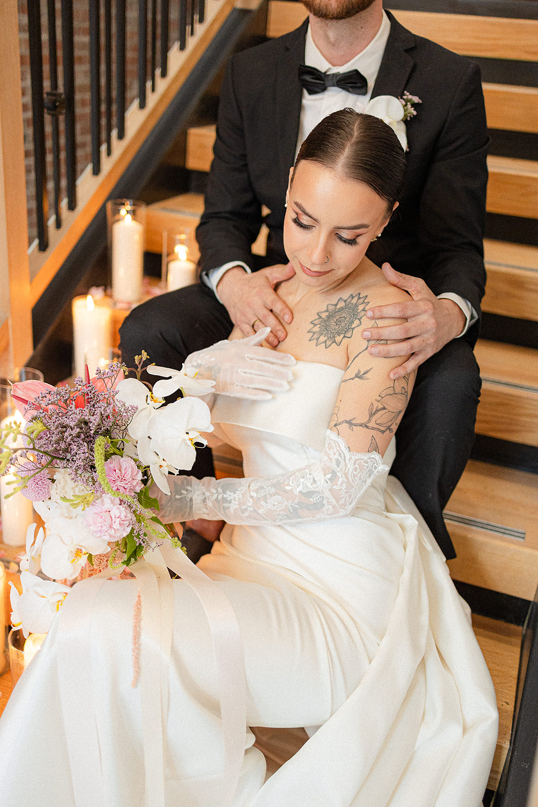 Bride wearing a white gown with lace gloves holding a floral bouquet, sitting on stairs with groom behind her in a black tuxedo.