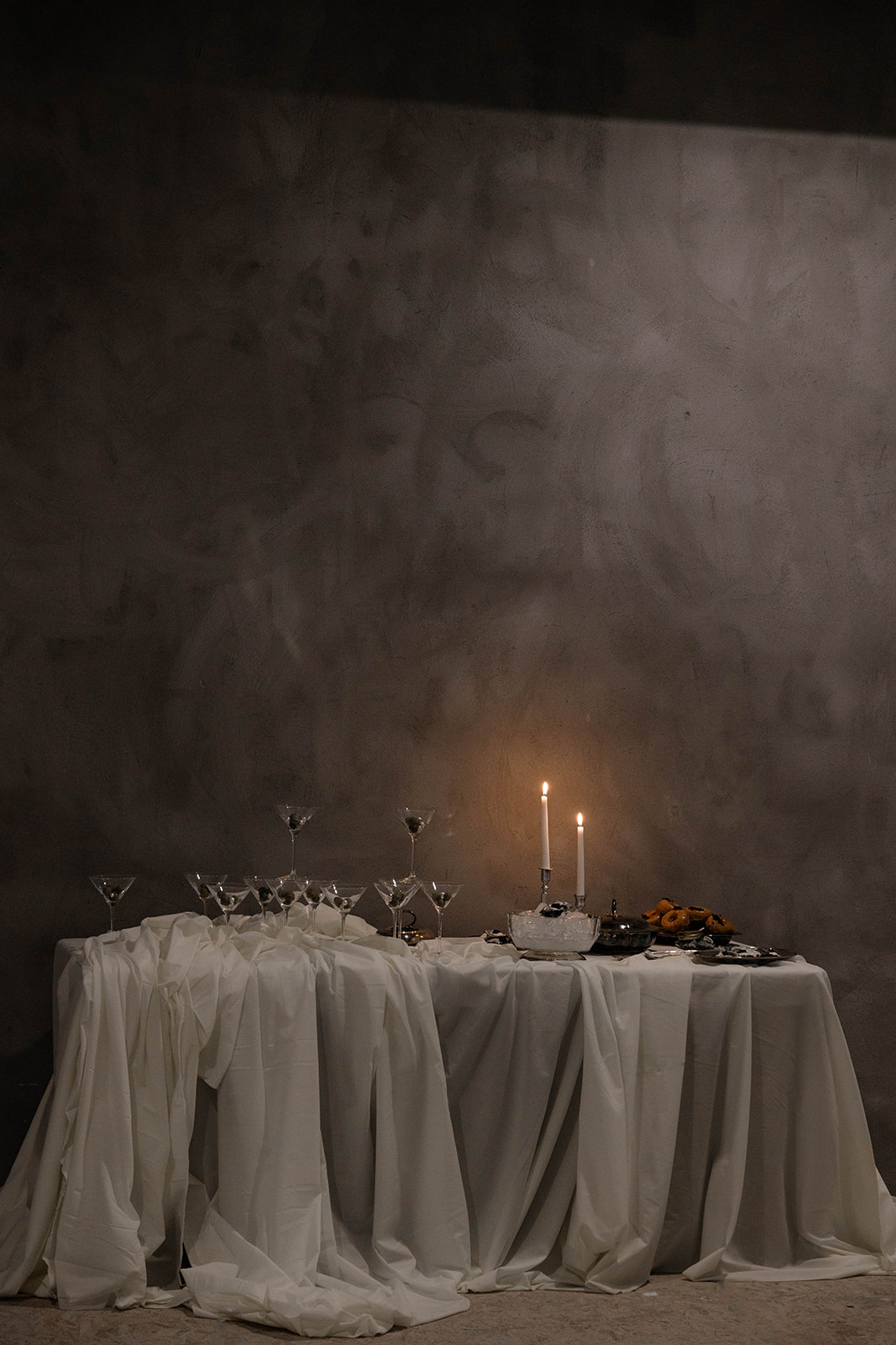 Table draped with white cloth holding martini glasses, two lit candles, and a plate of pastries against a dark textured wall.