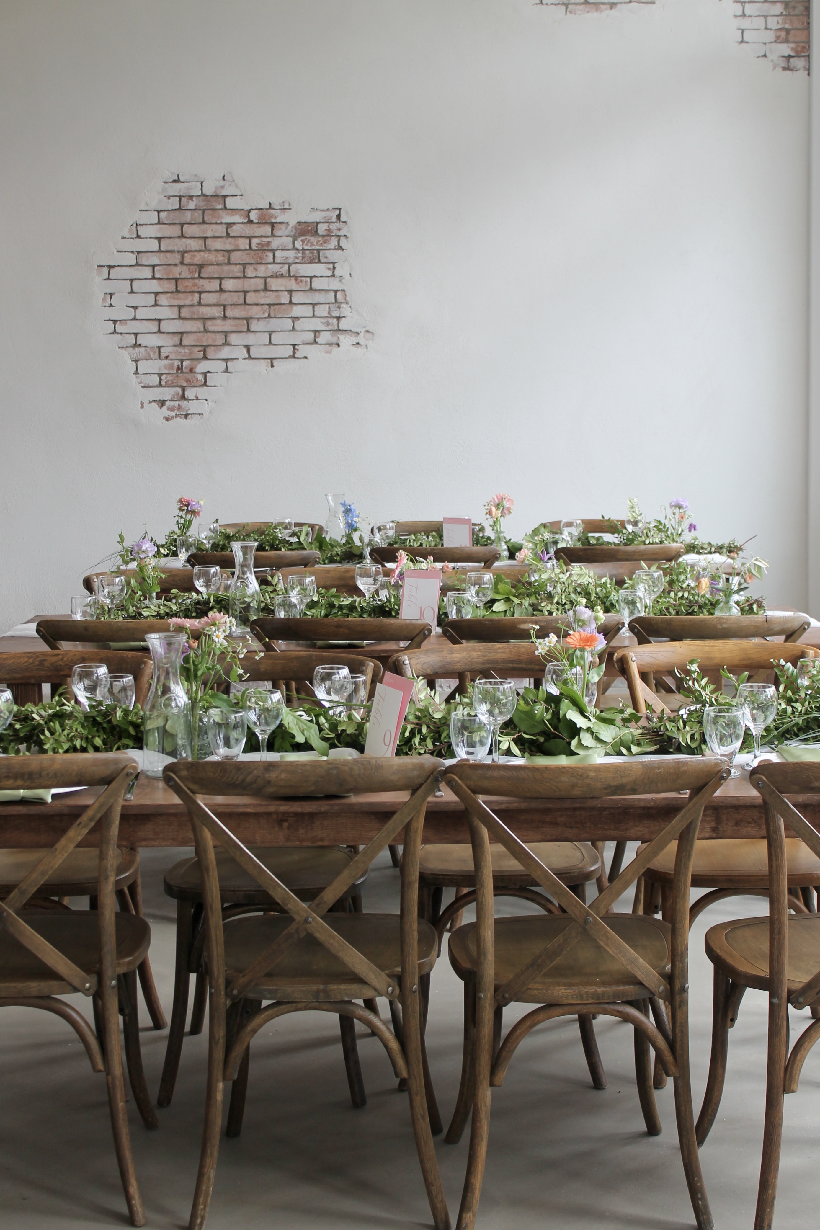 Long wooden farmhouse tables with rustic wooden chairs, decorated with greenery, flowers, glassware, and table number cards against a white wall with exposed brick patches.