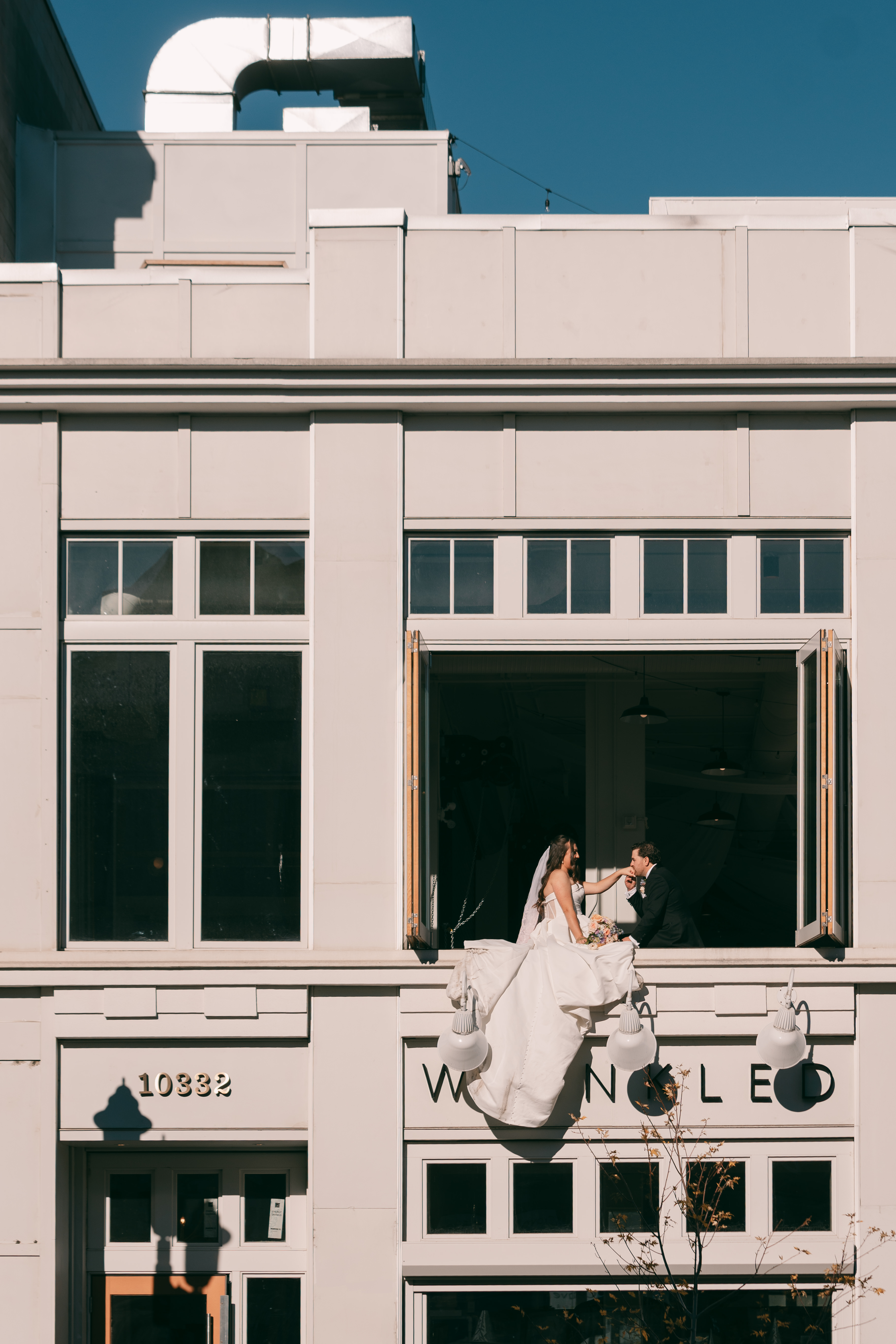 Bride in white dress and groom in black suit sitting on a ledge of an open window on a white building.