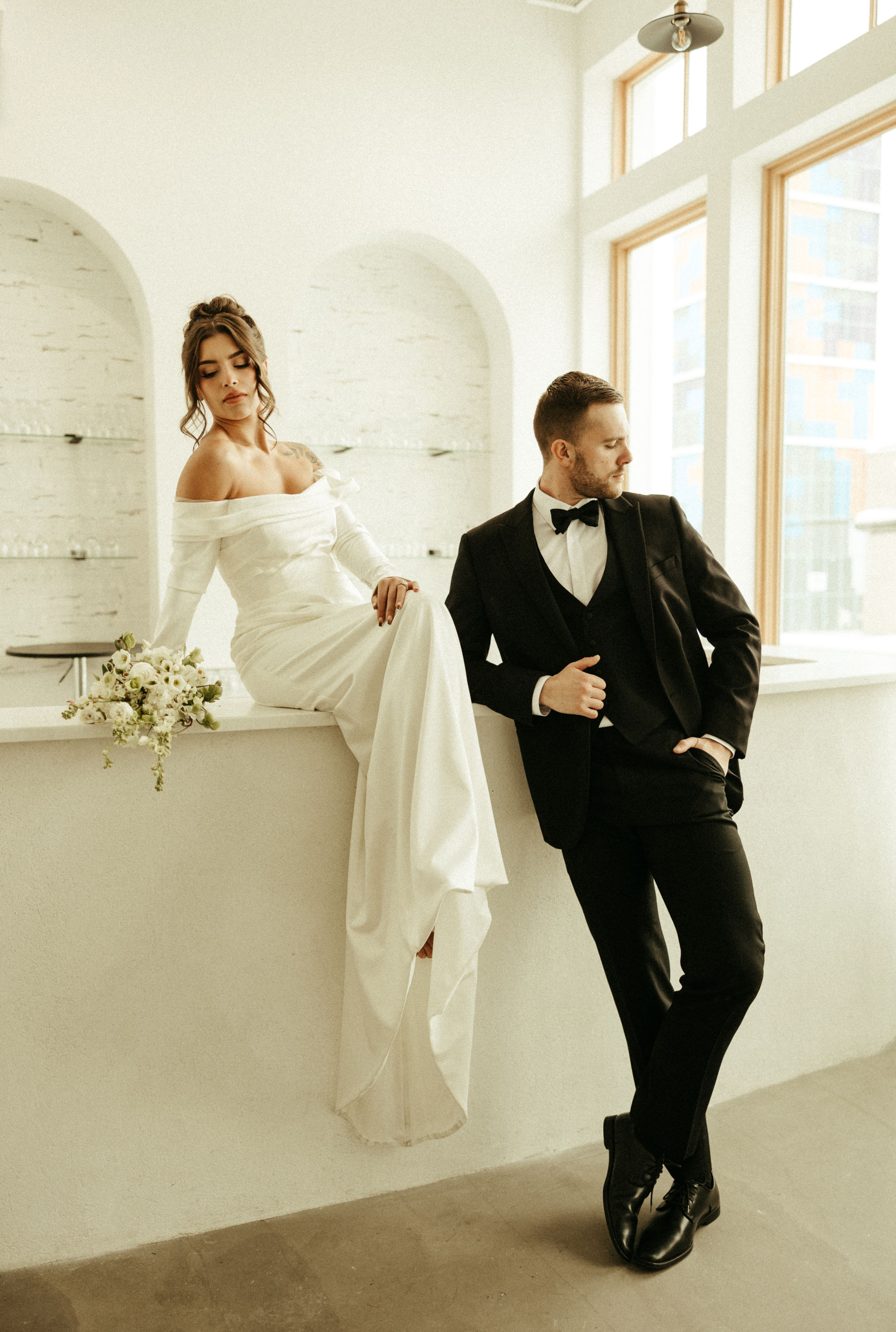 Bride in an off-shoulder white gown sitting on a counter holding a bouquet, groom in a black suit leaning against the counter in a modern bright room.