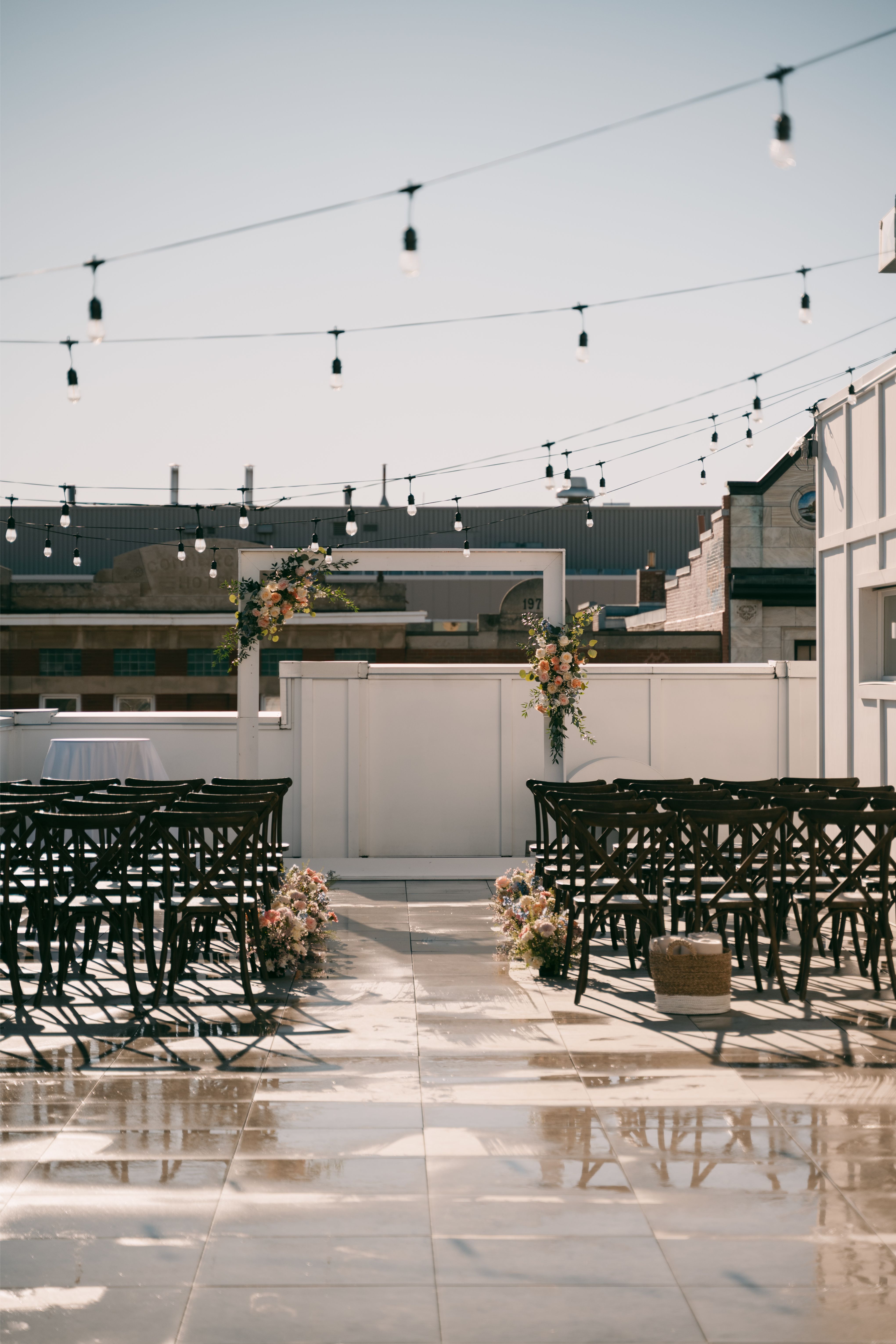 Rooftop wedding ceremony setup with wooden chairs, floral decorations, string lights, and a white arch.