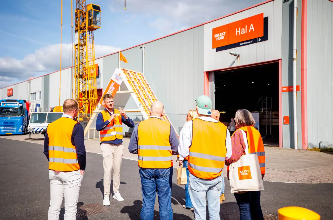 Groepje mensen staat met een oranje hesje voor een prefabhal