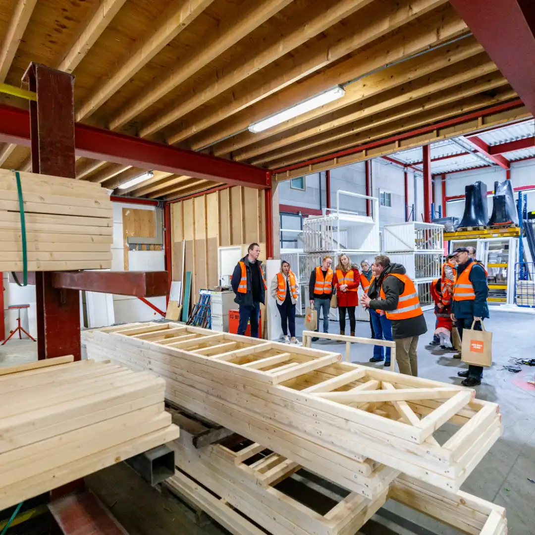 Een groep mensen met oranje hesjes staat in een prefabhal