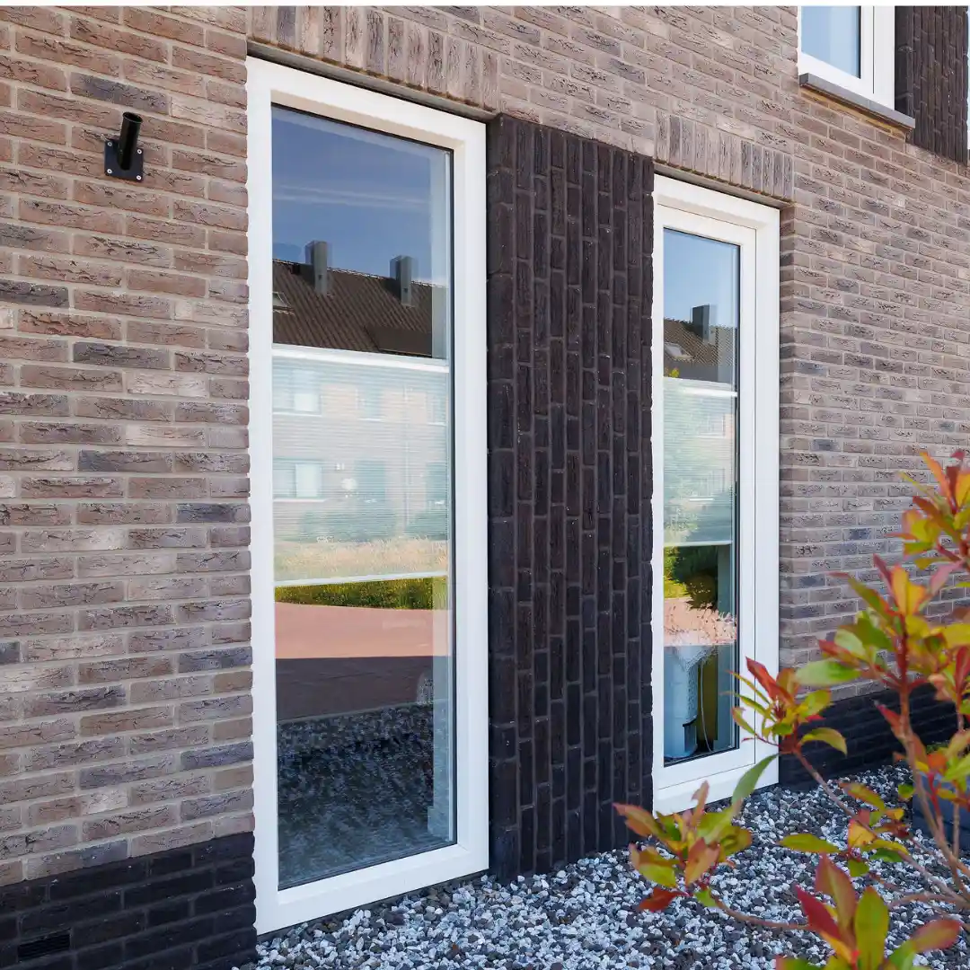 Twee grote ramen aan de zijkant van de woning met rechtopstaande donkere stenen in het midden.