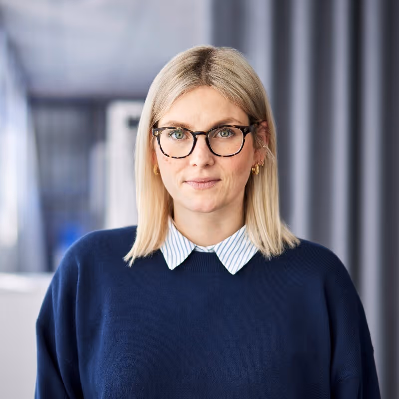 Frau mit blonden Haaren, die eine dunkelblaue Pullover und eine gestreifte Hemdbluse mit Kragen trägt, sowie eine Brille mit dunklem Rahmen und goldene Ohrringe.