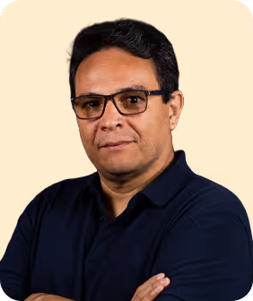 A man with short black hair and glasses, wearing a navy polo shirt, poses with arms crossed against a light beige background.