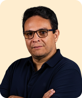 A man with short black hair and glasses, wearing a navy polo shirt, poses with arms crossed against a light beige background.