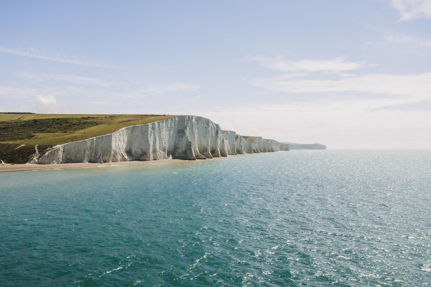 White chalk cliffs rise from turquoise sea with green grassland above.