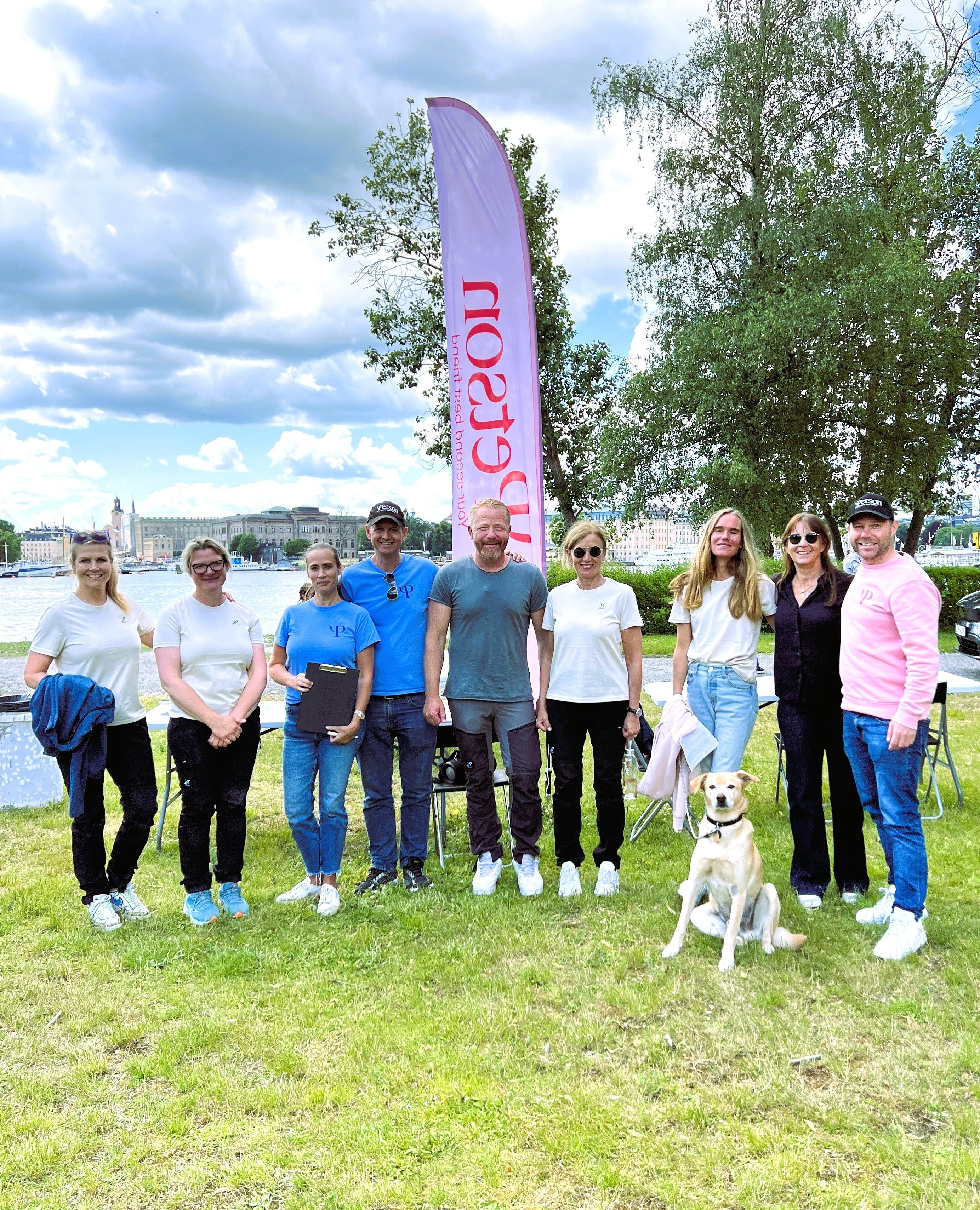 Group of nine people standing on grass near water with trees and city buildings in the background, a dog sitting in front, and a tall Pets on flag behind them.