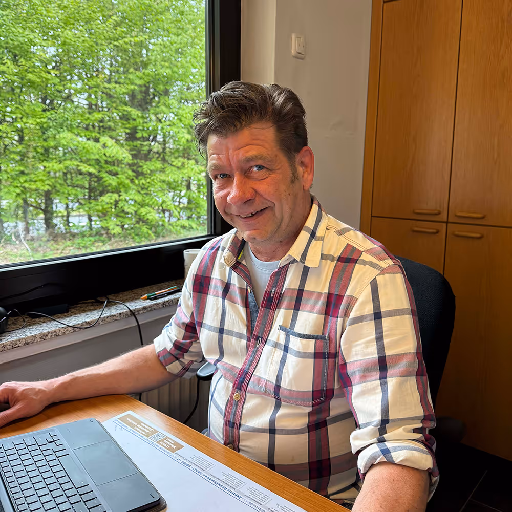 Smiling man in a plaid shirt sitting at a desk with a laptop next to a window showing green trees outside.