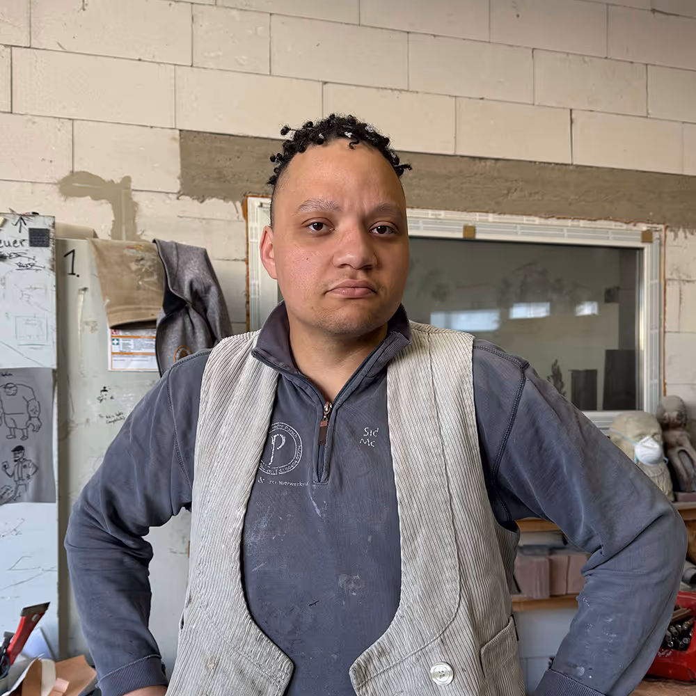 Man with short curly hair standing with hands on hips in a workshop with a gray shirt and white vest.