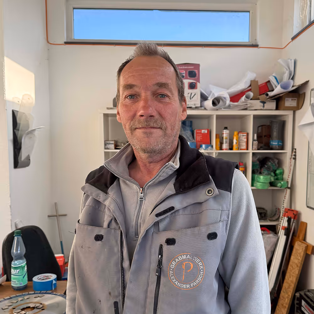 Man with short hair and light facial hair wearing a gray work vest with the logo 'Grabmalwerk Alexander Parsch' standing in a cluttered workshop.