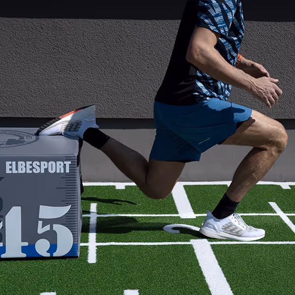 Ole Nauert, Inhaber und Therapeut der Rheinblick Physio, zeigt Split Squats mit Hilfe einer Plyo Box auf dem grünen Sprinter-Teppich im Außenbereich der Rheinblick Physio.