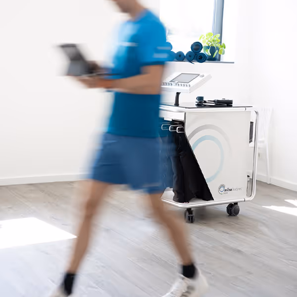 Das mobile EMS Gerät im Fokus, Ole Nauert, Inhaber und Therapeut der Rheinblick Physio, unscharf zu sehen, läuft mit IPad in der Hand im Vordergrund durch den Raum.