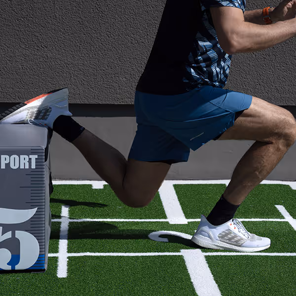 Ole Nauert, Inhaber und Therapeut der Rheinblick Physio, zeigt Split Squats mit Hilfe einer Plyo Box auf dem grünen Sprinter-Teppich im Außenbereich der Rheinblick Physio.