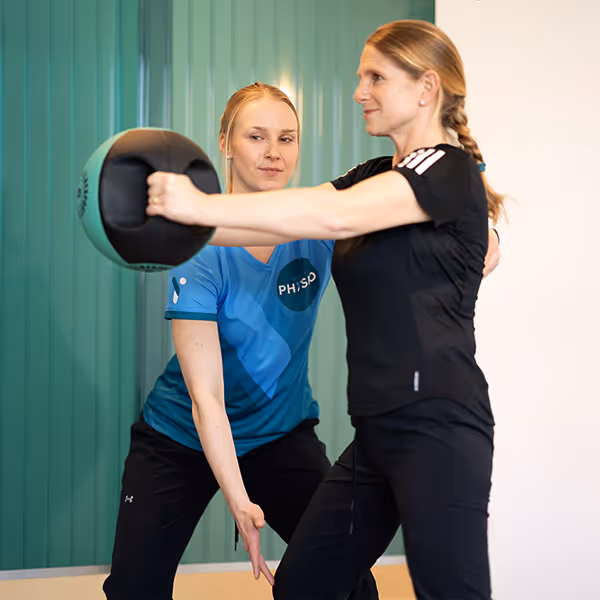 Eine Patientin der Rheinblick Physio macht eine Rückenübung mit Hilfe eines Medizinballs. Dabei wird ihre Haltung von einer Therapeutin korrigiert.