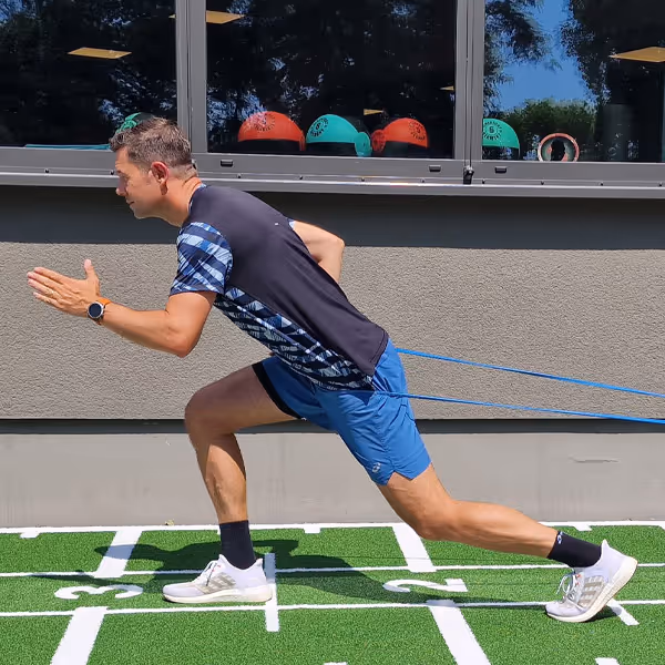 Ole Nauert, Inhaber und Therapeut der Rheinblick Physio, zeigt im Außengelände auf dem grünen Sprinter-Teppich eine Sprintübung gegen ein Widerstandsband.